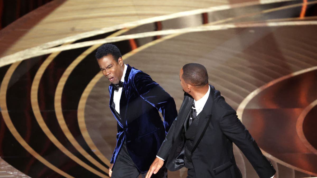 Will Smith in a black suit strikes Chris Rock in a blue velvet suit while on stage presenting at the Oscars