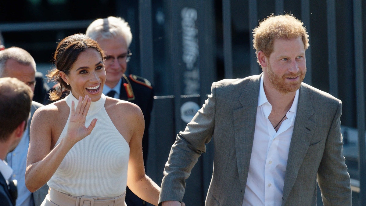 Meghan Markle waves in a white halter top and light pink pants while holding Prince Harry's hand, who sports a gray suit