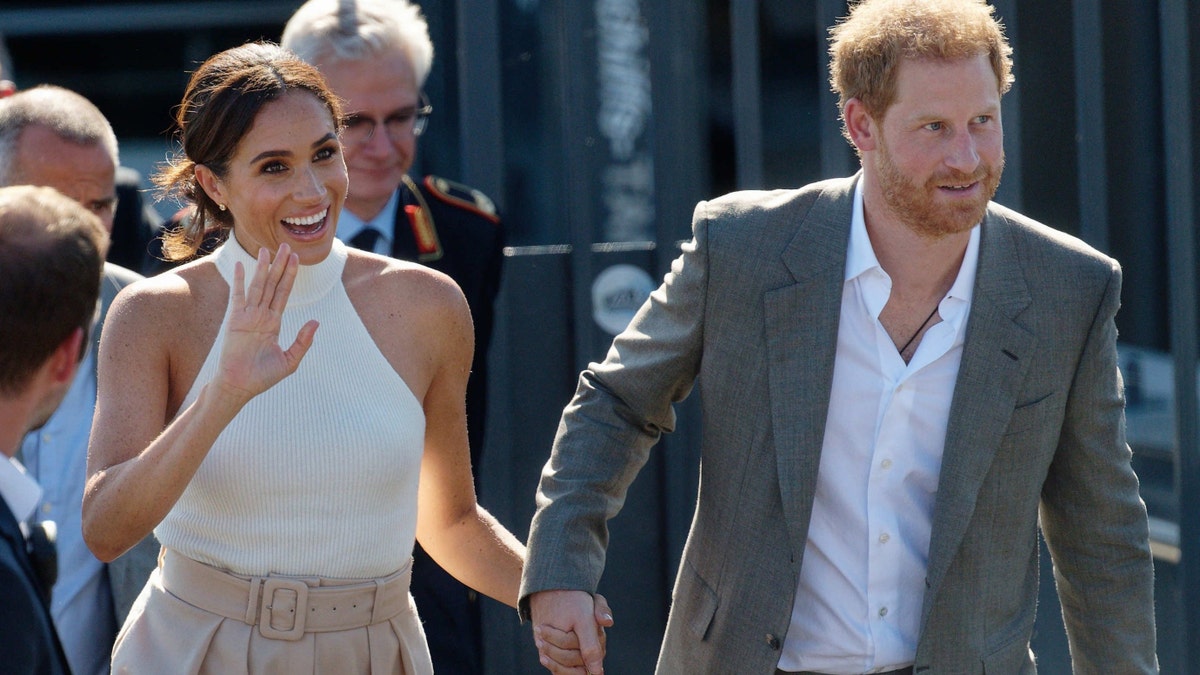 Meghan Markle waves in a white halter top and light pink pants while holding Prince Harrys hand in a grey suit