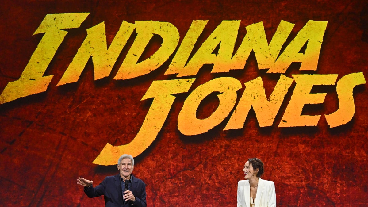 Harrison Ford and Phoebe Waller-Bridge speak to a crowd in front of a large Indiana jones back drop with red coloring and yellow words