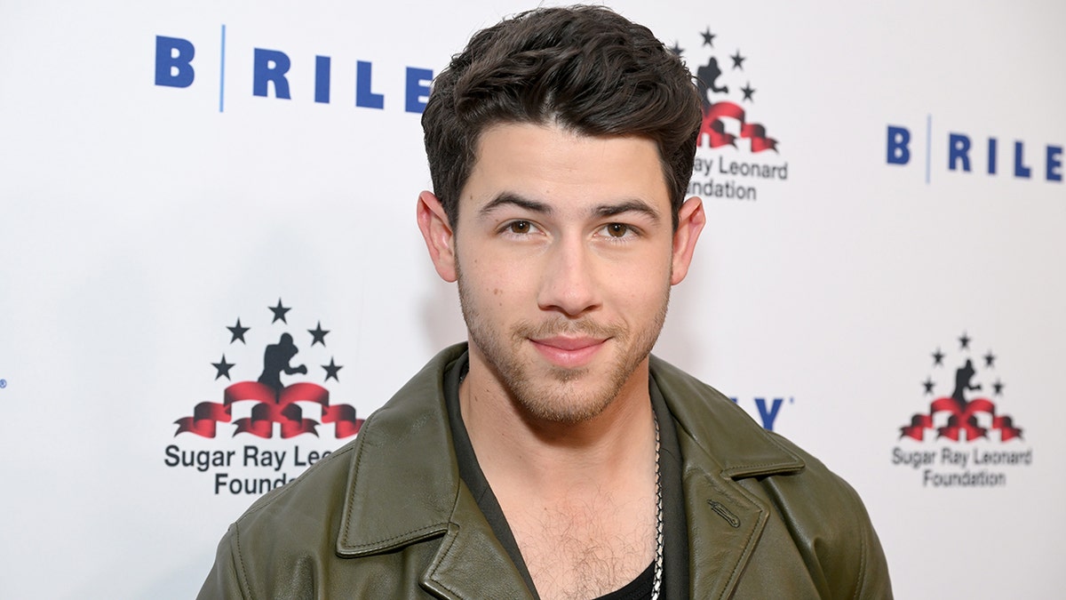 NIck Jonas smiles in an olive green coat at a Sugar Ray Leonard Foundation event