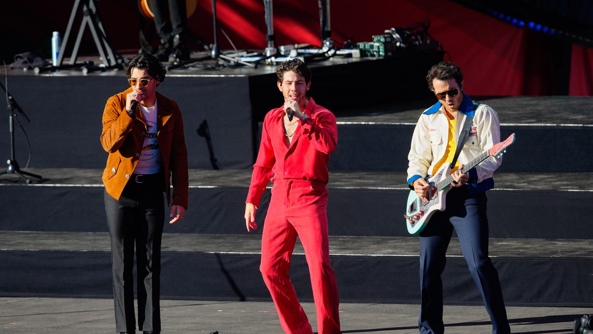 Joe in orange, Nick in red suit, and Kevin in a white and blue jacket while performing as the "Jonas Brothers"