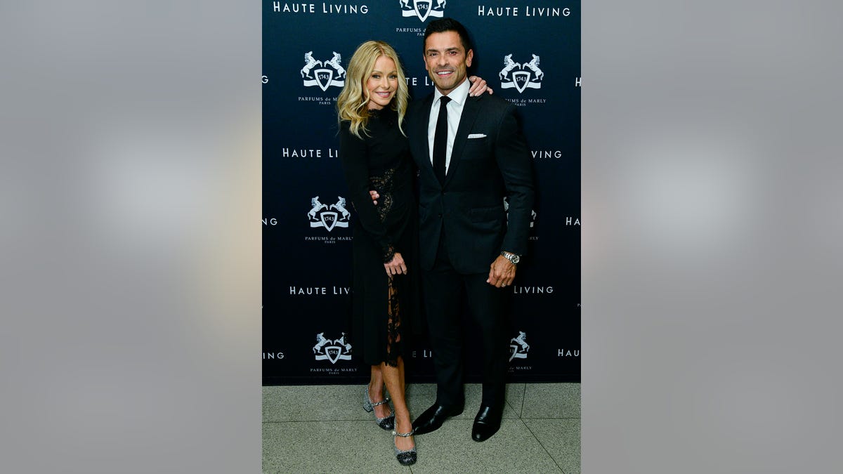 Kelly Ripa in a long black dress poses with her husband Mark Consuelos in a white shirt, black tie and black suit
