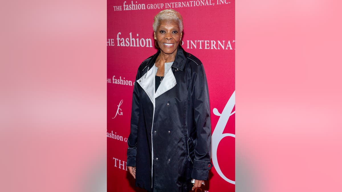 Dionne Warwick in a black coat with white lapels and a red background