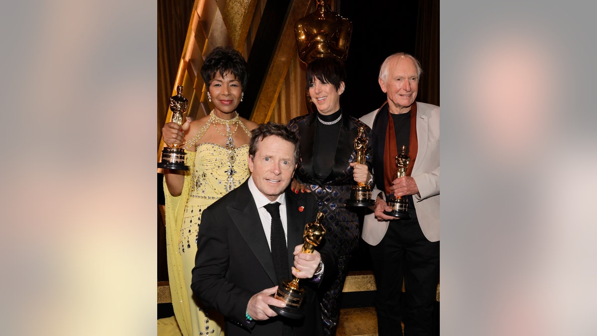 Euzhan Palcy in a yellow dress, Michael J. Fox in a black suit and tie, Diane Warren in all black, and Peter Weir in a grey suit and red shirt pose for a photo with their awards from the Governors Awards