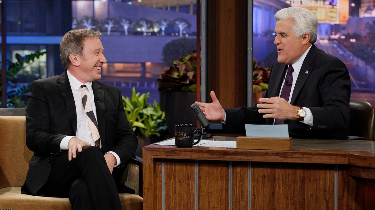 Tim Allen in a white shirt and black suit talks with Jay Leno behind the desk at The Tonight Show