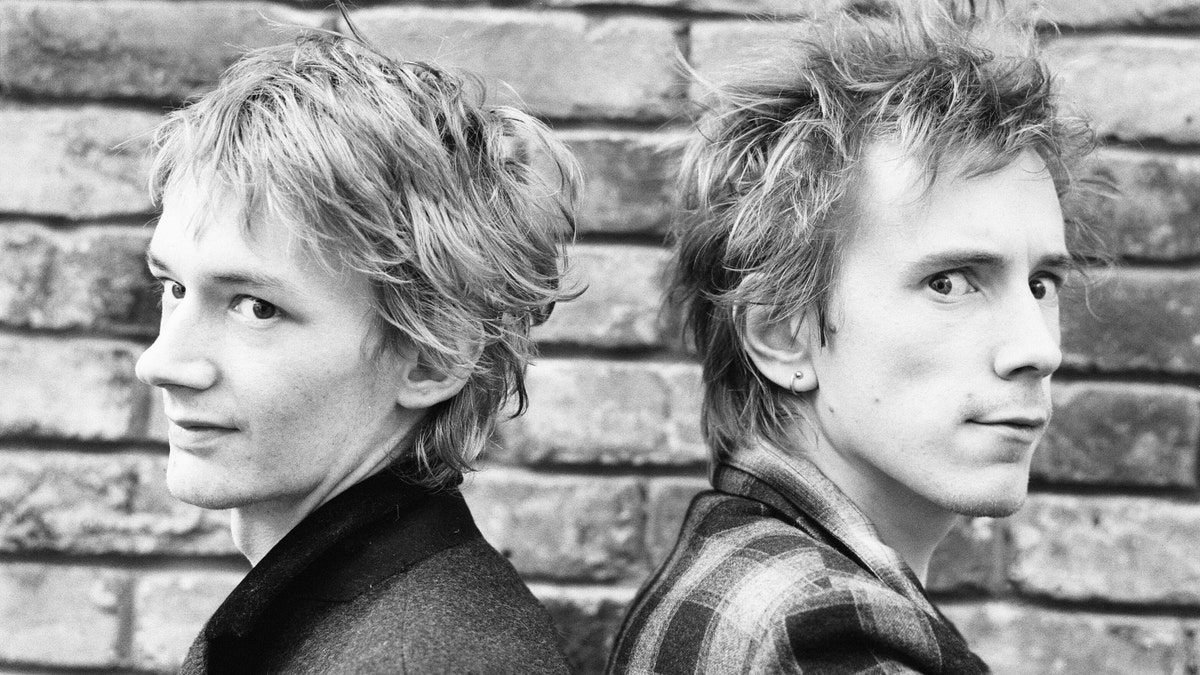 Johnny Rotten and Keith Levene stand pose in front of a brick wall in a black-and-white photo