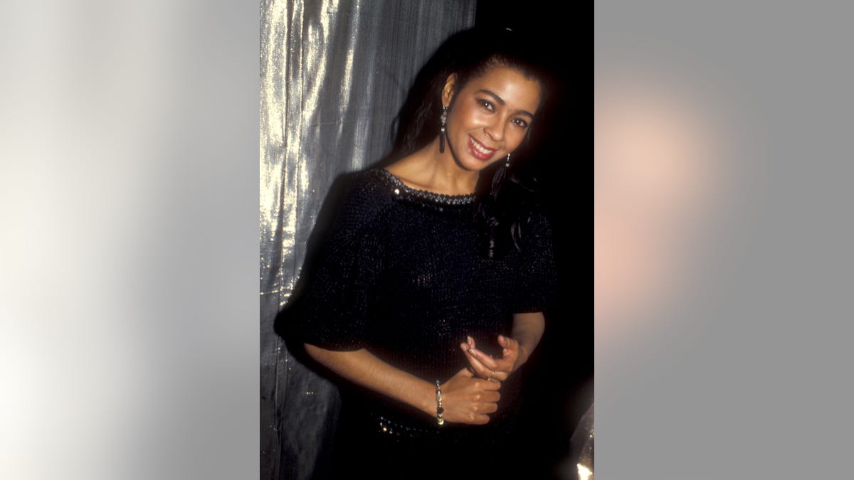 Irene Cara smiles for a photo in a black outfit and dangly earrings