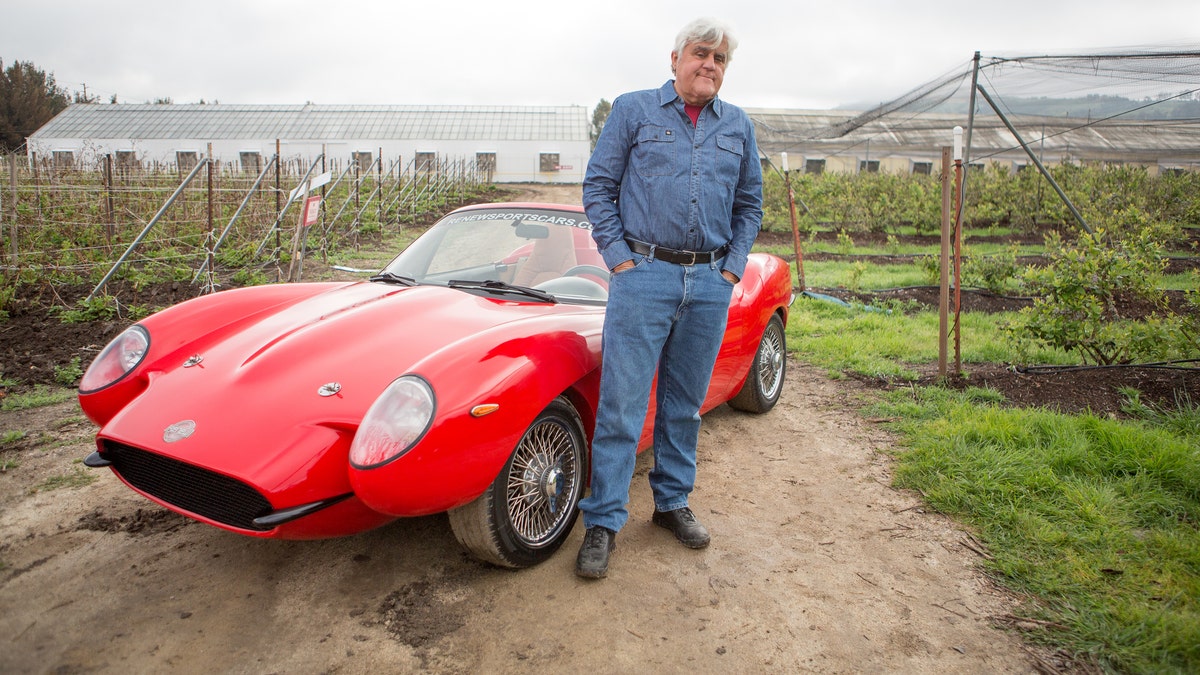Jay Leno poses with a red sports car for his show about rare vehicles