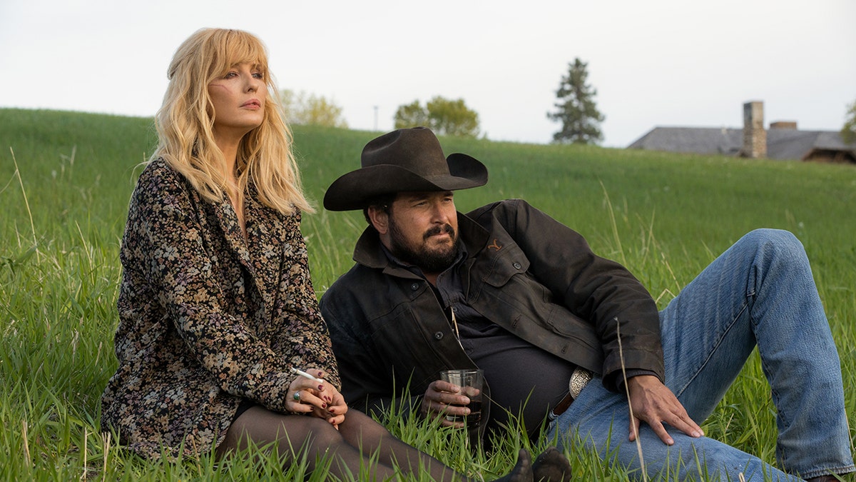 Cole Hauser as Rip Wheeler and Kelly Reilly as Beth Dutton sitting in the grass for a "Yellowstone" stock picture