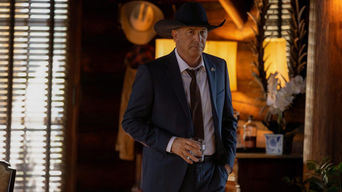 Kevin Costner holds a glass wearing a blue suit and dark brown cowboy hat as John Dutton in "Yellowstone"