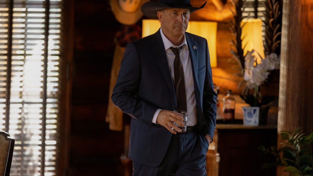 Kevin Costner holds a glass wearing a blue suit and dark brown cowboy hat as John Dutton in "Yellowstone"