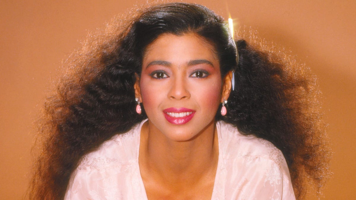 Irene Cara smiles in a portrait with a white dress and light pink earrings with an orange backdrop