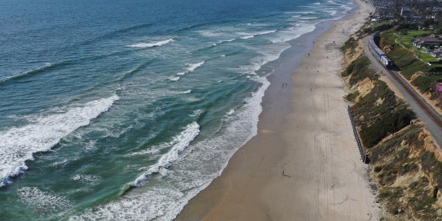 An Amtrak Pacific Surfliner train passes in front of pedestrians walking along a beach in Del Mar, California, on May 13, 2021.