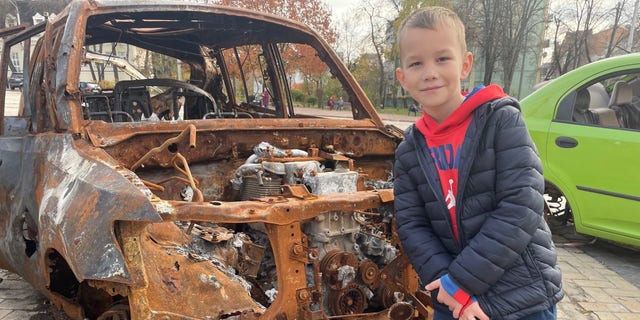 A young boy in Kyiv standing in front of a bombed out car.