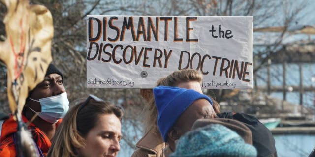 A man holds a sign that reads "Dismantling the Doctrine of Discovery" as people march during the National Day of Mourning on Thanksgiving Day, November 25, 2021, in Plymouth, Massachusetts.