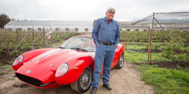 Jay Leno poses with a red sports car for his show about rare vehicles