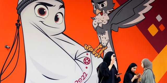 Women stand in front of a billboard showing an illustration of the upcoming Qatar 2022 FIFA World Cup mascot