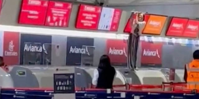 Otros pasajeros en el aeropuerto pueden ver a la persona fuera de control, de pie en el mostrador de facturación y sosteniendo una pantalla sobre ella, desde lejos.