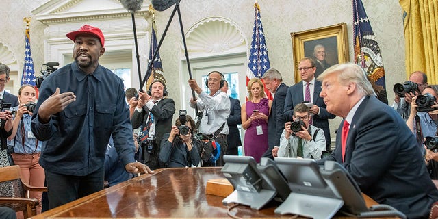 Surrounded by members of the press and others, American rapper and producer Kanye West stands as he talks with real estate developer and  then-President Donald Trump in the White House's Oval Office, Washington DC, October 11, 2018. West wears a red baseball cap that reads 'Make America Great Again,' Trump's campaign slogan. 