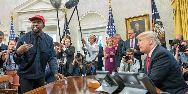 Surrounded by members of the press and others, American rapper and producer Kanye West stands as he talks with real estate developer and  then-President Donald Trump in the White House's Oval Office, Washington DC, October 11, 2018. West wears a red baseball cap that reads 'Make America Great Again,' Trump's campaign slogan. 