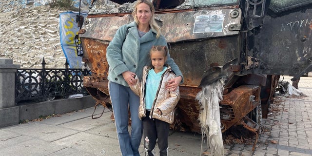 A mother and daughter in Kyiv stand in front of a damaged military vehicle.