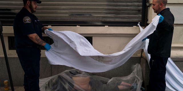 Forensic assistant Laurentiu Bigu, left, and investigator Ryan Parraz from the Los Angeles County coroner's office cover the body of a homeless man found dead from overdose on a sidewalk on April 18, 2022.