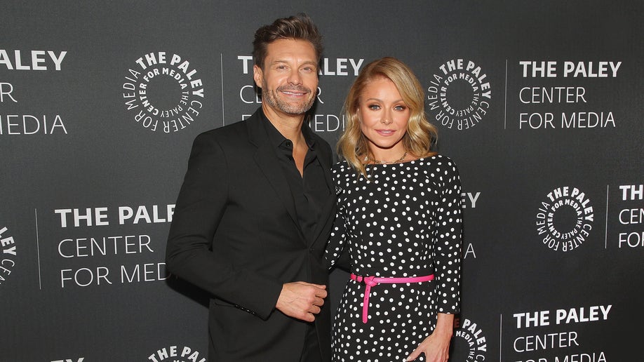 Ryan Seacrest in a black suit poses alongside Kelly Ripa in a black polka dot dress with a pink ribbon