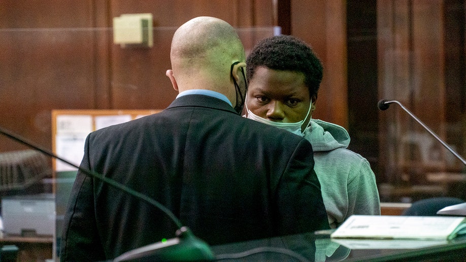 lawyer with his back to the camera, speaks to defendant who is dressed in a grey hooded sweatshirt and wears a medical mask