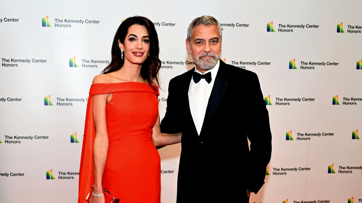 Amal Clooney in a off-the-shoulder-red gown with a large red cape poses on the red carpet with husband George Clooney in a classic tuxedo