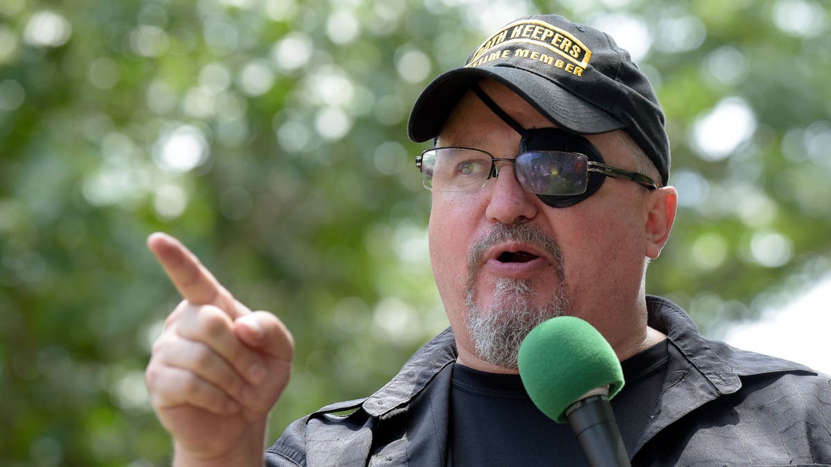 Stewart Rhodes with an eye patch, holding a microphone and pointing while giving a speech