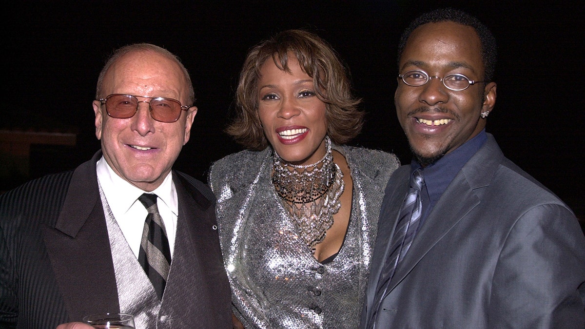 Clive Davis in a black suit stands next to Whitney Houston in a shimmery silver suit who stands next to Bobby Brown in a navy suit