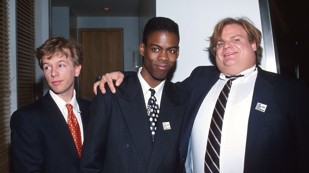 David Spade in a suit and red tie looks off camera while posing with Chris Rock in a dark suit and tie who is hugged by Chris Farley