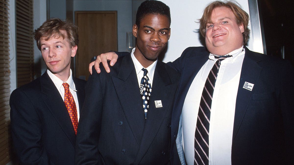 David Spade in a suit and red tie looks off camera while posing with Chris Rock in a dark suit and tie who is hugged by Chris Farley