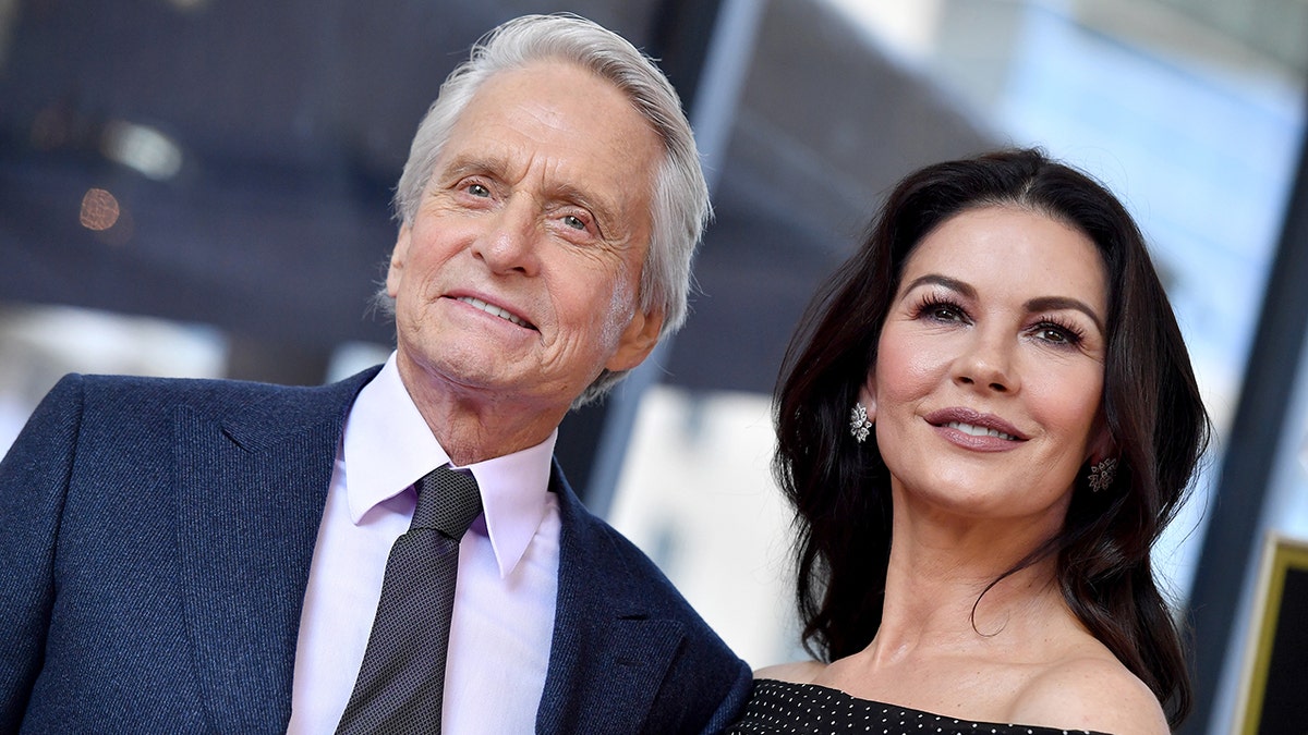 Michael Douglas in a white shirt and navy blazer with a tie smiles off in the distance with wife Catherine Zeta-Jones in a black and white polka dot dress