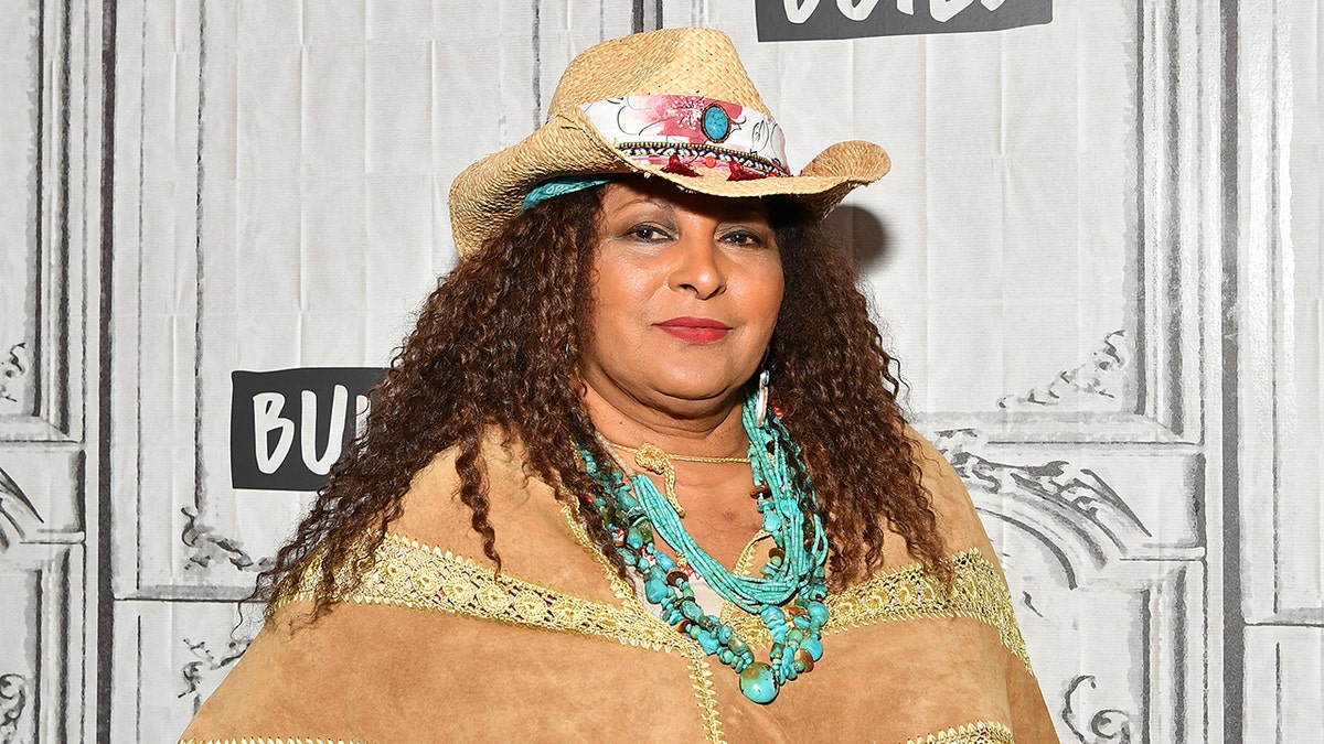 Pam Grier in a tan hat and matching tan suede top and a turquoise chunky necklace