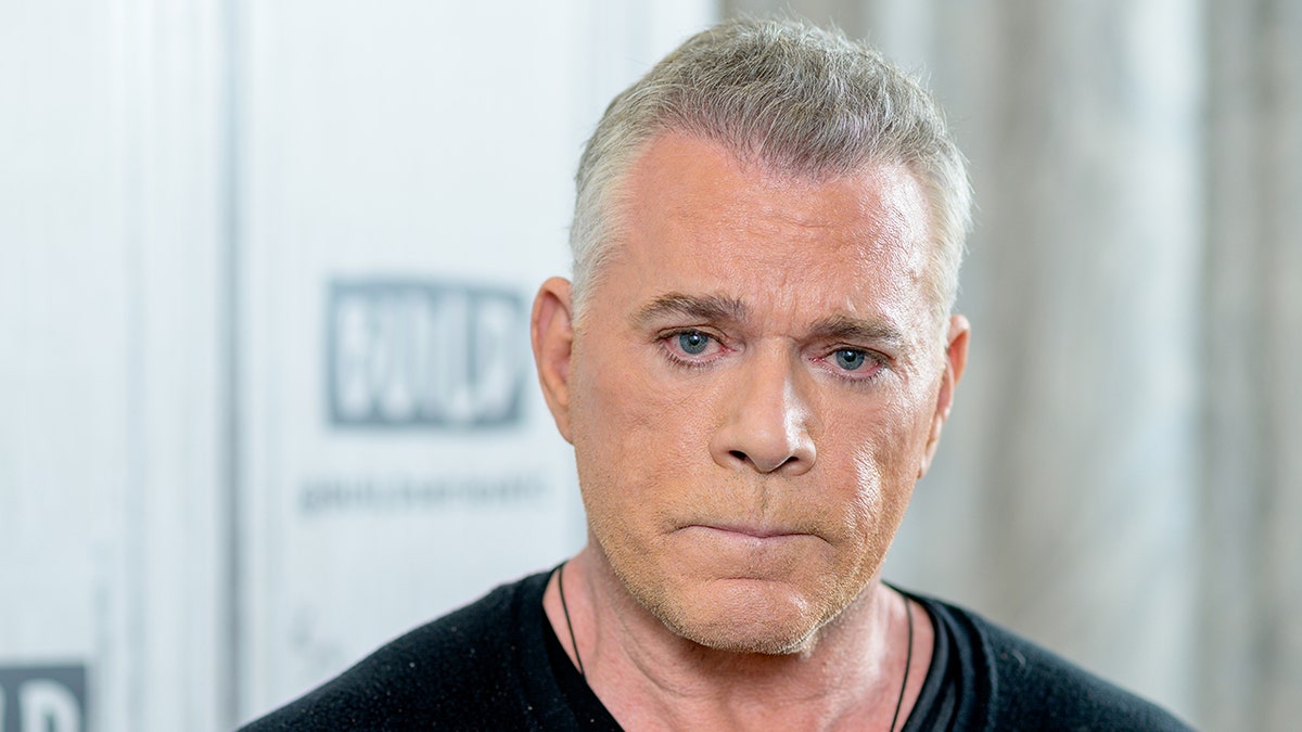 Ray Liotta in a black shirt and chain necklace looks directly at the camera