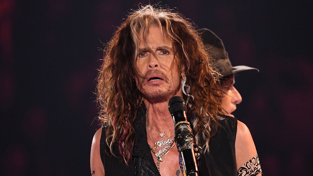 Steven Tyler on stage in front o a microphone in a black low cut top
