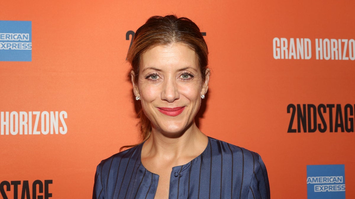 Kate Walsh smiles in a blue low cut striped dress against an orange background