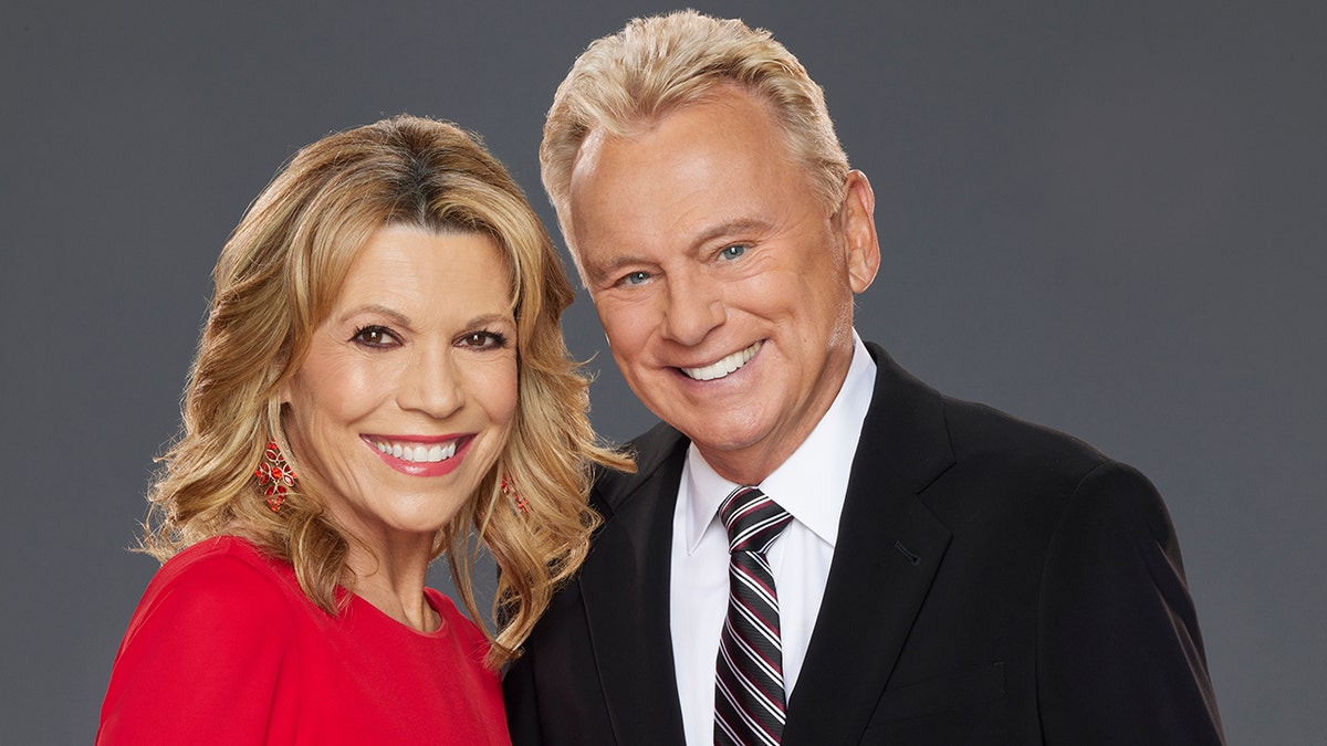 Vanna White in a red dress poses with Pat Sajak in a black suit and patterned tie for "Celebrity Wheel of Fortune" promo shots