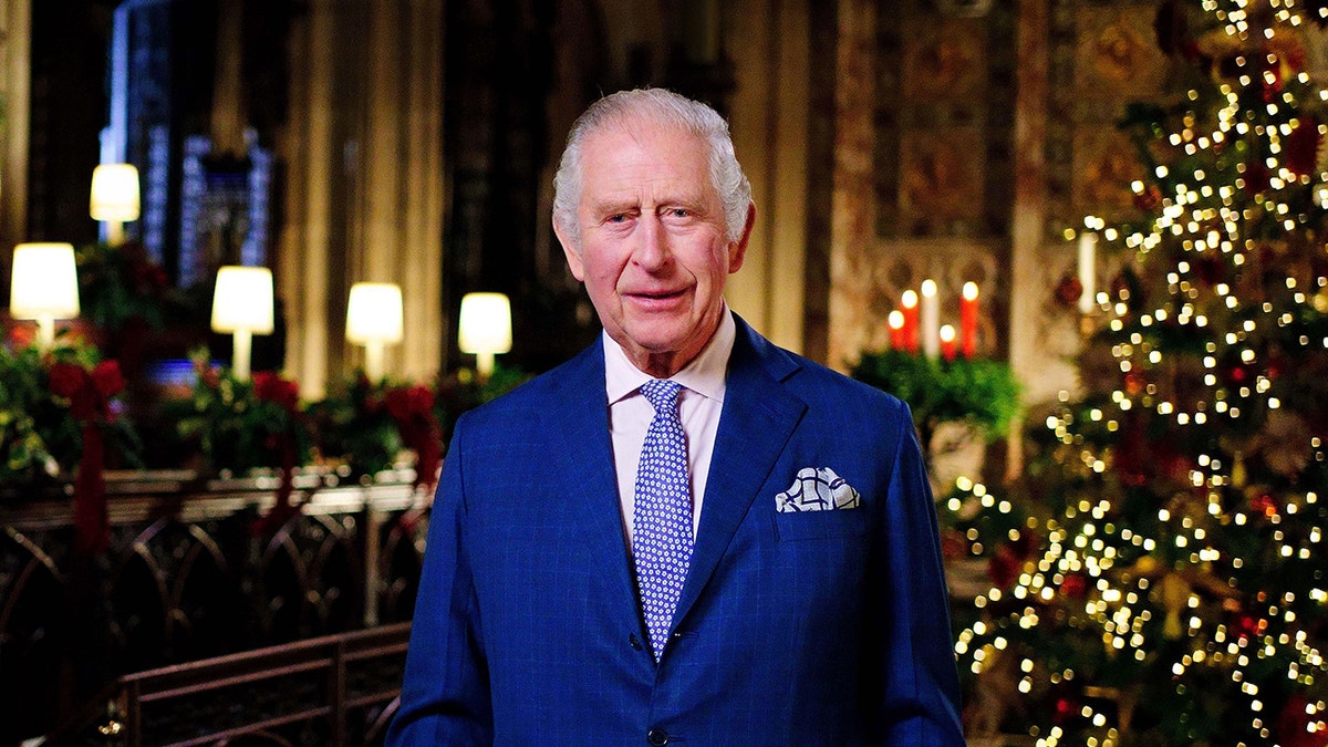 King Charles in a royal blue suit and light blue tie stands in front of a lighted Christmas tree and lit candles to give his speech