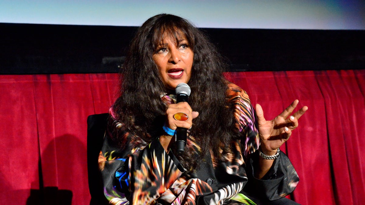 Pam Grier in a patterned black top on stage speaking into a microphone