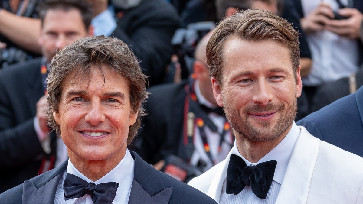 Tom Cruise in a black tuxedo smiles alongside co-star Glen Powell in a white tuxedo on the red carpet in Cannes, France
