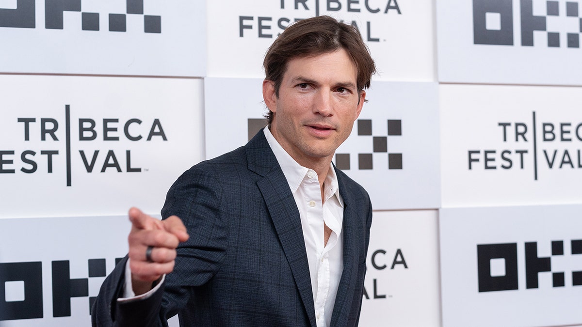 Ashton Kutcher points on the red carpet in a crisp white shirt and blue suit blazer