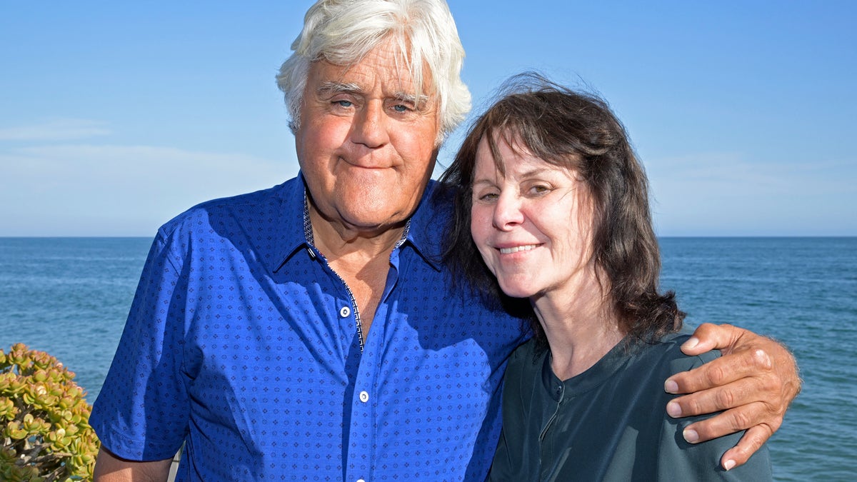 Jay Leno in a royal blue button down wraps his arm around wife Mavis in a dark teal shirt