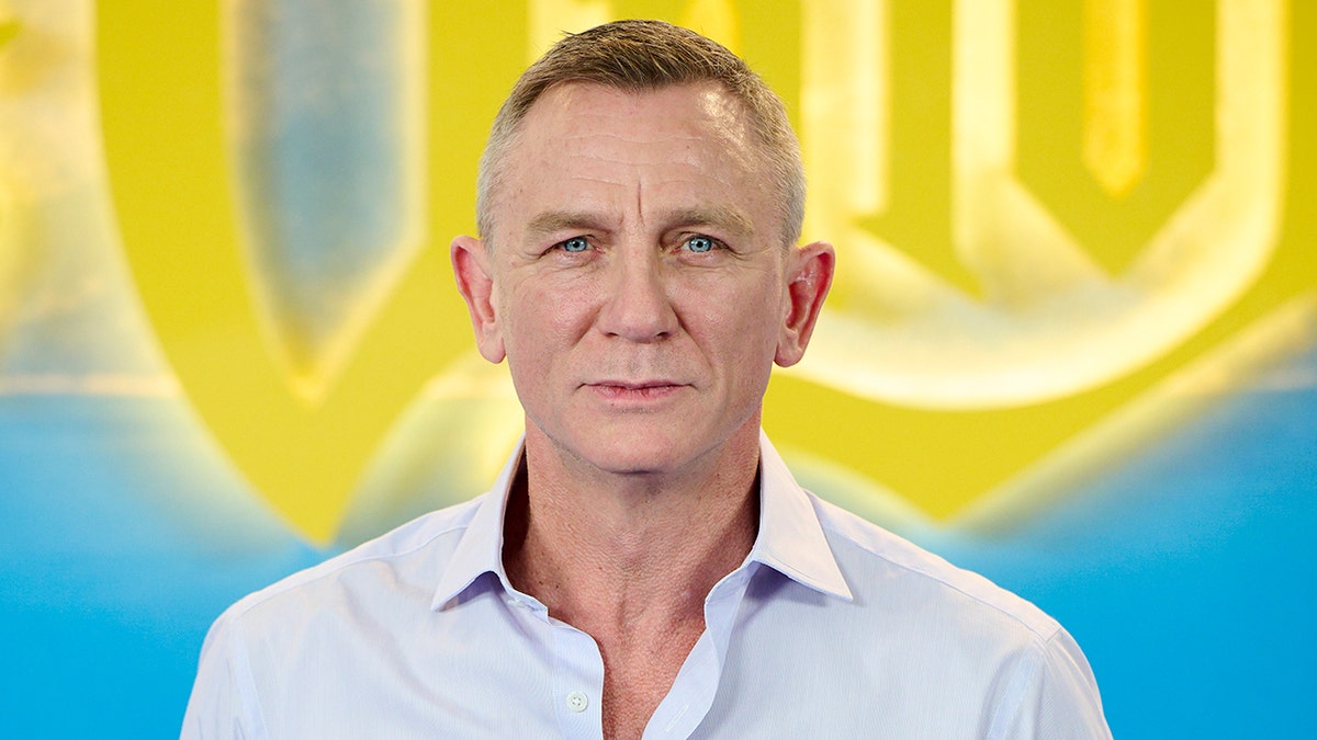 Daniel Craig in a very light blue button down shirt in front of a bright yellow and blue background for "Glass Onion: A Knives Out Mystery"