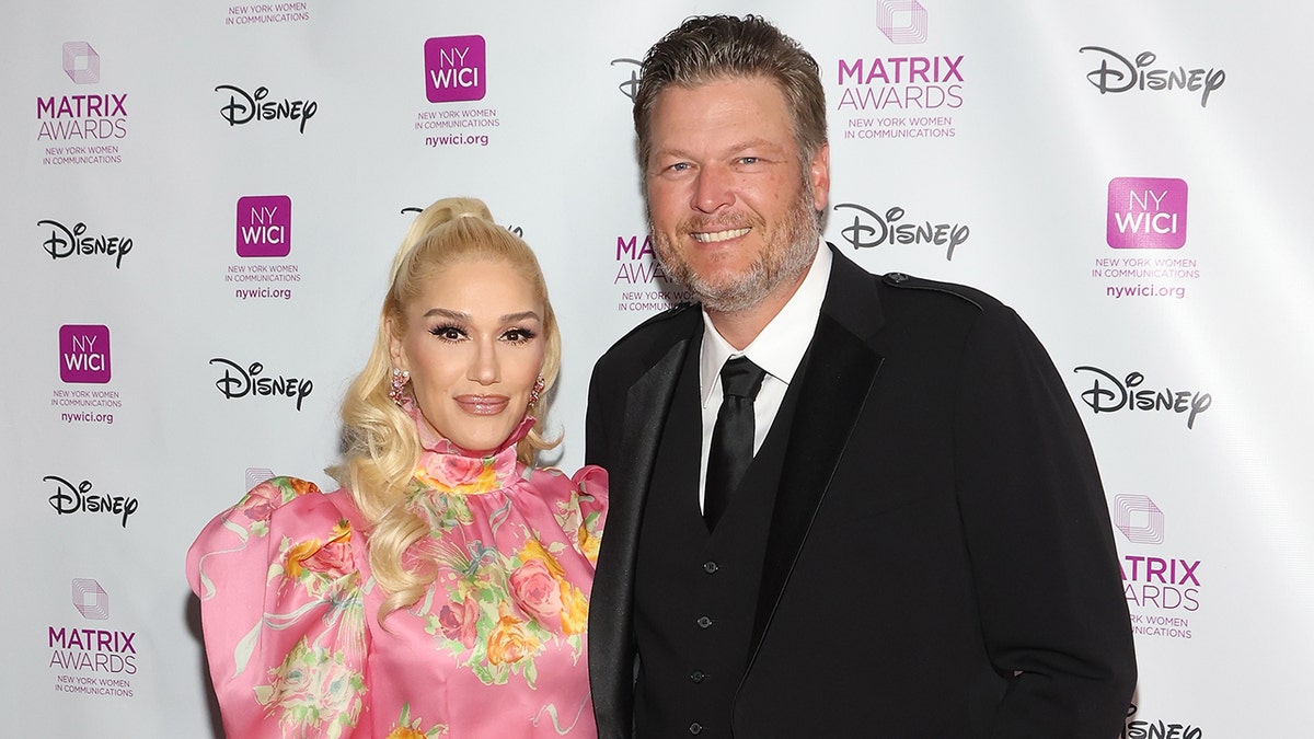 Blake Shelton in a black suit and black tie poses with Gwen Stefani in a bubblegum pink floral dress with poof shoulders