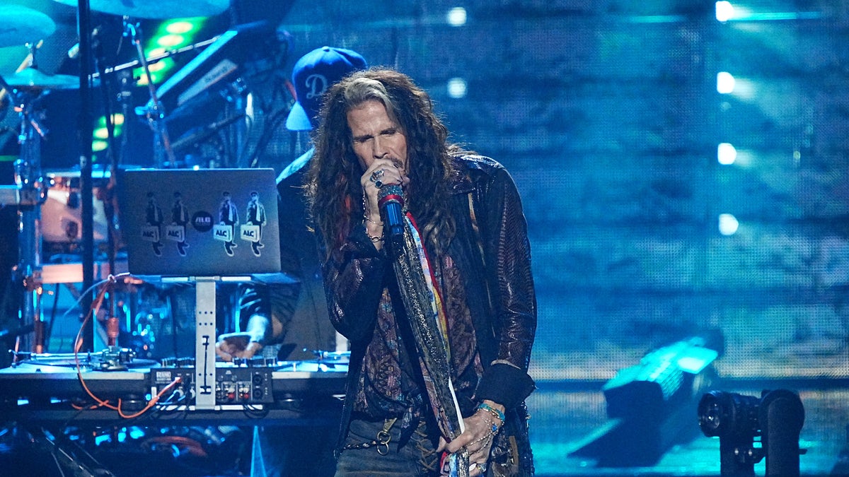 Steven Tyler in jeans and black leather jacket sings into the microphone