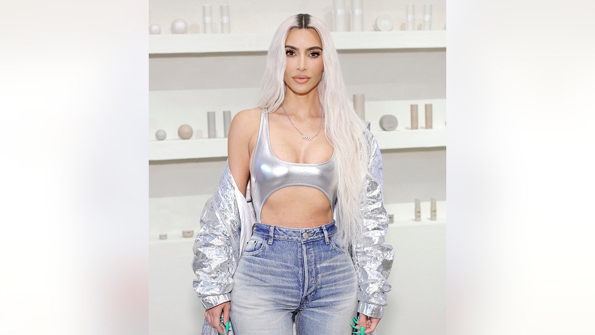 Kim with platinum long blonde hair in a cut-out silver spandex body suit and jeans and a silver jacket off her shoulders