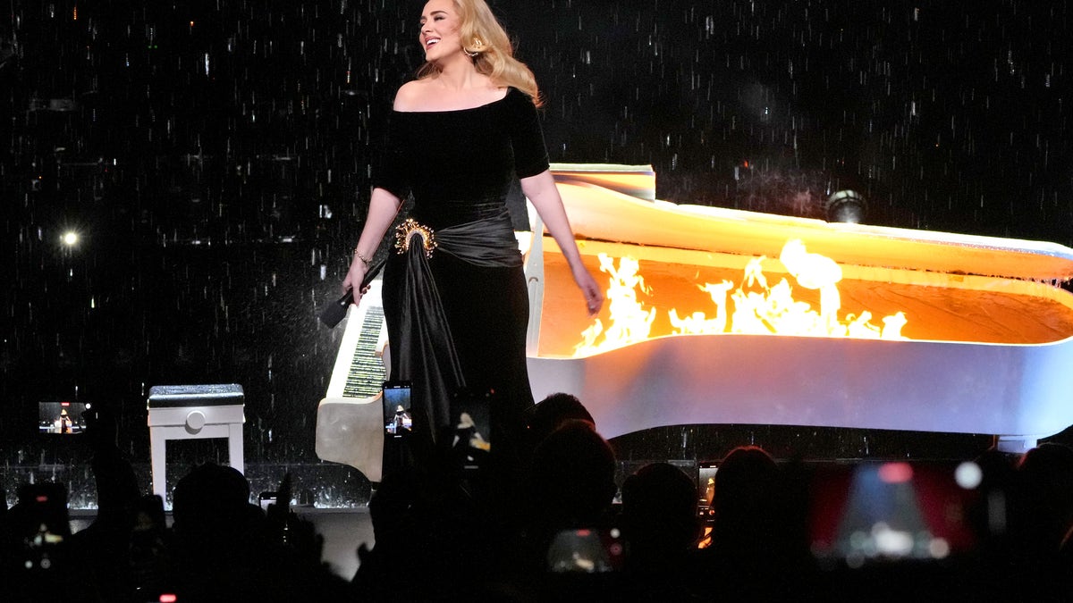Adele in a black outfit with a white piano up in flames behind her during her concert